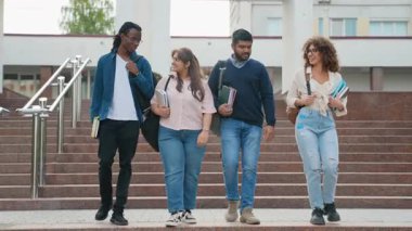 Kitaplar ve sırt çantaları taşıyan dört farklı üniversite öğrencisi modern bir kampüs binasının merdivenlerinden birlikte yürüyor, konuşuyor ve gülümsüyor, arkadaşlığı ve yüksek eğitimi temsil ediyor.