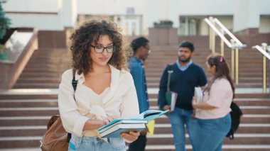Merdivenlerde duran gözlüklü ve sırt çantalı genç bir kız öğrencinin portresi yukarı bakıp gülümsemeden önce arka planda konuşan diğer uluslararası öğrencilerle birlikte bir kitap okuyor.