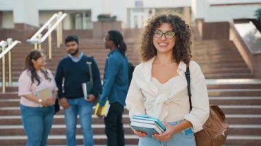 Kıvırcık saçlı ve gözlüklü genç üniversite öğrencisi elinde kitaplarla kampüs merdivenlerinde gülümserken çeşitli sınıf arkadaşları arka planda, modern akademik hayatta sohbet ediyorlar.