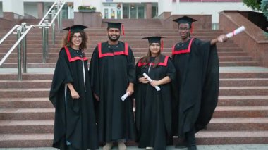 Siyahlar içinde dört mutlu üniversite mezunu merdivenlerde durmuş akademik başarılarını kutlamak için gülümsüyor ve diplomalarını yükseltiyorlar.