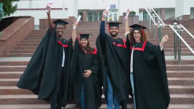 Üniversite kampüsünün merdivenlerinde akademik önlükler ve şapkalarla kutlama yapan, diplomalarını yükselten ve kameraya birlikte gülümseyen çok ırklı yüksek lisans öğrencileri grubu.