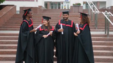 Üniversite merdivenlerinde diplomaları olan, gülümseyen, başarıyı, dostluğu ve birlikte parlak bir geleceği kutlayan, akademik elbiseler ve havan mermileri içinde dört mutlu çok ırklı mezun.