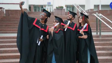 Çeşitli gülümseyen mezunlar üniversite basamaklarında, kolej basamaklarında havan merdiveni ve elbiseler giyerek başarılarını ve dostluklarını yakalamak için selfie çekiyorlar.