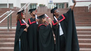 Mezuniyet elbisesi içinde üniversite kampüsünün merdivenlerinde akıllı bir telefonla selfie çekerek akademik başarılarını kutlayan çok ırklı bir grup öğrenci.