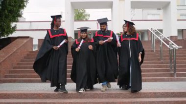 Çeşitli çok kültürlü akademik mezunlar üniversite merdivenlerinde yürüyorlar, diplomalarını tutuyorlar ve mezuniyet töreninden sonra birlikte konuşuyorlar.
