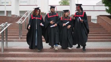 Geleneksel akademik şapkalar ve cübbeler içinde dört gülümseyen etnik üniversite mezunu kampüs binasının dışındaki merdivenlerden birlikte inerek başarılarını kutluyorlar.