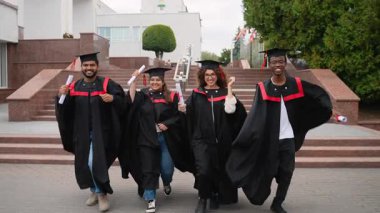 Heyecanlı çok ırklı yüksek lisans öğrencileri akademik önlük ve şapkalar içinde birlikte yürüyorlar, başarılarını kutluyorlar ve mezuniyet töreninden sonra diplomalarını tutuyorlar.