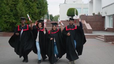 Siyahlar içinde çok uluslu mutlu mezunlar üniversite kampüsünde yürüyorlar, akademik başarılarını gururla kucaklayarak ve sarmalanmış diplomalarını sallayarak kutluyorlar.