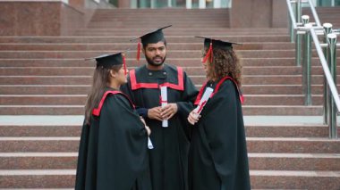 Siyahlar içinde üç mutlu üniversite mezunu ellerinde diplomalarla üniversitenin merdivenlerinde durup gelecekleri hakkında konuşarak başarılarını kutluyorlar.