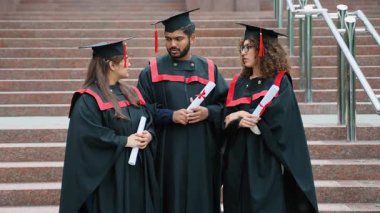 Mezuniyet elbisesi ve şapkalı üç genç uluslararası öğrenci ellerinde diplomalarıyla üniversitenin merdivenlerinde canlı bir konuşma yapıyorlar.