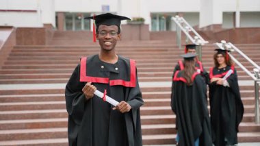 Kameraya gururla gülümseyen kep ve cüppeli genç Afro-Amerikan erkek mezun. Diploma tutuyor. Üniversite merdivenlerinde arka planda kutlama yapan bir grup farklı öğrenci var.