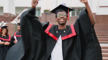 Mezuniyet elbisesi ve şapkası içinde gururlu Afro-Amerikan erkek öğrenci, başarısını bir diplomayla kutluyor, sevinçle sınıf arkadaşlarıyla el kaldırıyor kampüs merdivenlerinde arka planda.