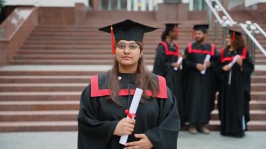 Geleneksel kep ve cübbeyle mezun olan genç Hintli kız üniversite diplomasını gururla tutarken aynı zamanda gülümsüyor ve çeşitli öğrenci arkadaşları arka planda ayakta duruyor.