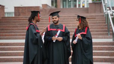 Üç mutlu çok kültürlü üniversite mezunu şapkalı ve cüppeli merdivenlerde diplomalarını tutuyorlar. Gülümseyen uluslararası öğrenciler akademik başarılarını birlikte kutluyorlar.