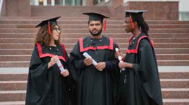 Geleneksel akademik önlüklü ve şapkalı üç farklı üniversite öğrencisi ellerinde diplomalarıyla kampüs merdivenlerinde durmuş başarılı mezuniyet törenlerini tartışıyorlar.