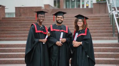 Üç mutlu çok ırklı üniversite öğrencisi, akademik önlük ve şapkalarla merdivenlerde, diplomalarını tutuyorlar ve kampüsteki mezuniyet törenlerinden sonra kameraya gülümsüyorlar.