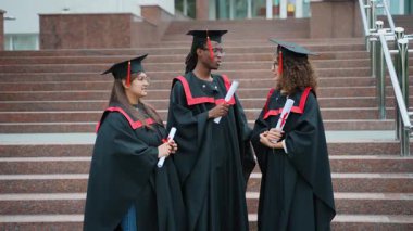 Geleneksel akademik şapkalı ve cüppeli üç mutlu üniversite öğrencisi kampüs binasının dışındaki merdivenlerde duruyorlar.