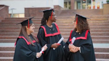 Üç mutlu çok ırklı üniversite mezunu, akademik önlük ve şapkalarla merdivenlerde durmuş, diplomalarını tutuyor ve kampüsteki mezuniyet töreninden sonra neşeli bir şekilde sohbet ediyorlar.
