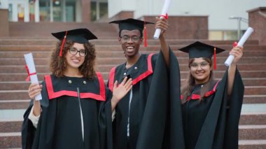 Üç üniversite mezunundan oluşan mutlu bir grup akademik elbiseler ve havan topları içinde gülümseyip, diplomalarını kampüs binasının merdivenlerinde gururla sallıyor ve kutluyorlar.