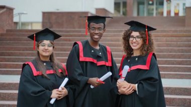 Üç farklı ve mutlu üniversite mezunu, geleneksel akademik kepler ve cübbeler giyerek diplomalarını alıp törenden sonra gururla gülümsüyorlar.