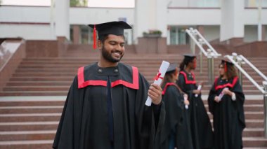 Mezuniyet gününde akademik başarısını kutlamak için üniversite basamaklarında diplomasını tutarken gururla gülümseyen elbisesi ve havan topu giymiş mutlu Hintli öğrenci.