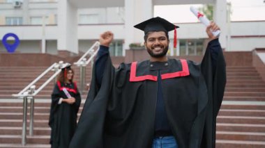 Akademik başarısını kutlamak için geleneksel bir elbise ve havan tahtasıyla başarılı bir Hintli mezun, gururla bir diploma tutuyor ve üniversite merdivenlerinde kollarını kaldırıyor.