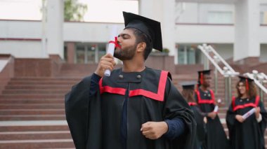 Geleneksel bir elbise ve havan topu şapkasıyla gururlu Hintli mezun diplomasını öpüyor, akademik başarısını ve başarısını dışarıda üniversite binasının önünde kutluyor.