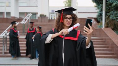 Mezuniyet elbisesi içindeki mutlu genç bir kadın, diplomasıyla akıllı bir telefonla selfie çekiyor, gülümsüyor ve arka planda sınıf arkadaşları dururken akademik başarısını kutluyor.