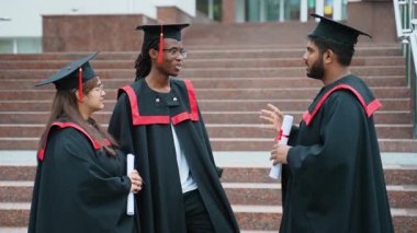 Geleneksel önlükler ve şapkalar giyen üç çok ırklı mezun üniversite merdivenlerinde ellerinde diplomalarla mezuniyetten sonra gelecekleri hakkında animasyon konuşuyorlar.