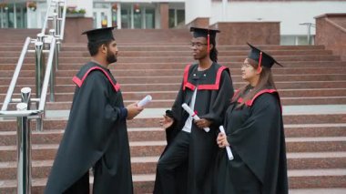 Mezuniyet elbisesi ve şapkası giyen üç farklı üniversite öğrencisi, mezuniyet töreninden sonra birlikte kampüs merdivenlerinde sevinçle konuşarak başarılarını kutluyorlar.