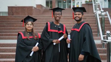 Akademik önlükler ve şapkalar içinde üç mutlu çok kültürlü mezun üniversite merdivenlerinde akademik başarılarını gururla kutluyor diplomalarını sallıyor ve kameraya gülümsüyor.