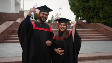 Başarılı bir çift akademik elbise içinde başarılarını kutlayan, diplomaları elinde tutarken mutlu bir şekilde gülümseyen ve törenden sonra üniversite kampüsünün merdivenlerinde duran yüksek lisans öğrencileri.