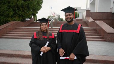 Mezuniyet töreninden sonra üniversite merdivenlerinde kırmızı kurdelelerle sarılı diplomalarını tutarak akademik elbiseler içinde gururlu Hintli mezunlar birlikte gülüyor ve başarılarını kutluyorlar.