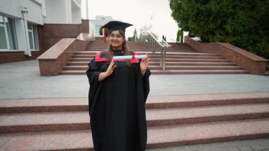 Mezuniyet gününde diploma tomarı tutarken geleneksel bir kep ve cübbe giyerek akademik başarısını kutlayan gururlu Hintli kadın mezun.