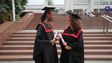 İki mutlu çok ırklı mezun, geleneksel siyah elbiseler ve şapkalar giyip, üniversite merdivenlerinde durmuş, birlikte çalışmalarının başarıyla tamamlanmasını kutluyorlar.