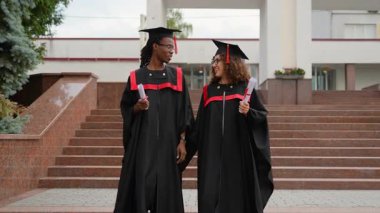 Diplomalarla kampüs merdivenlerinden inen akademik elbiseler içinde mutlu çok ırklı üniversite mezunları. Gururlu bir çift öğrenci başarılarının tadını çıkarıyor ve birlikte parlak geleceklerini kutluyorlar.