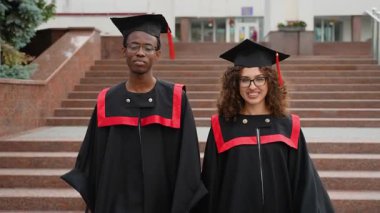 Gururlu çok ırklı bir çift, bir erkek ve bir kadın, üniversite merdivenlerinde mezuniyet elbiseleri ve şapkalarını takmış, törenden sonra birlikte gülümsüyorlar, başarılarını kutluyorlar.