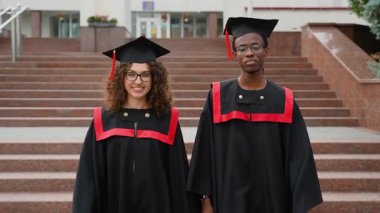 Geleneksel akademik şapkalar ve cüppelerle gülümseyen çok uluslu mezunlar, mezuniyetlerini üniversite merdivenlerinin önünde, kamera önünde gururla poz veriyorlar.