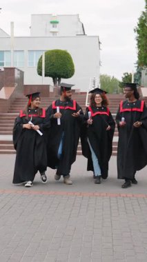 Mezuniyetten sonra üniversite kampüsünün yakınında birlikte yürüyüp diplomalarını ellerinde tutarak gülümseyen ve konuşan dört mutlu çok ırklı öğrenci.