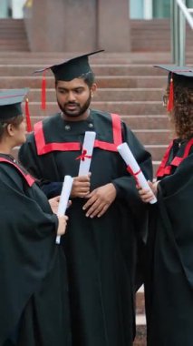 Mezuniyet töreninden sonra şapkalı ve cübbeli mutlu lisansüstü öğrencileri ellerinde diplomalarla kampüste keyifli bir sohbet ediyor ve akademik başarılarını kutluyorlar.