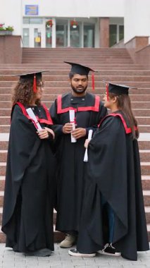 Siyahi akademik önlüklü ve şapkalı üç mutlu uluslararası öğrenci üniversitenin merdivenlerinde duruyor. Mezuniyet töreninden sonra diplomalarını alıp kampüsün önünde sohbet ediyorlar.