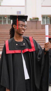 Geleneksel kep ve cübbeyle gülümseyen Afro-Amerikan erkek mezun seremoniden sonra diplomasını gururla tutuyor, akademik başarısını ve başarılı eğitimini kutluyor.