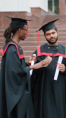 Geleneksel kep ve cüppe giymiş iki çok kültürlü erkek üniversite kampüsünün merdivenlerinde mezuniyet töreninden sonra mutlu mesut konuşuyorlar ve diplomalarını tutuyorlar.