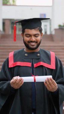 Geleneksel elbisesi ve akademik şapkası içinde kırmızı kurdeleli diploma tomarı tutan gururlu Hintli mezun öğrenci. Genç adam üniversite mezuniyetini ve eğitim başarısını kutluyor.