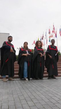 Çeşitli mutlu üniversite mezunları başarılarını uluslararası bayraklı bir binanın önüne havan topu kapaklarını atarak kutluyorlar.