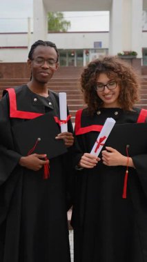 Diploma giyen ve kameraya gülümseyen iki mutlu çok ırklı öğrenci üniversite kampüsünün merdivenlerinde birlikte mezuniyet törenlerini konfetiler düşerek kutluyorlar.