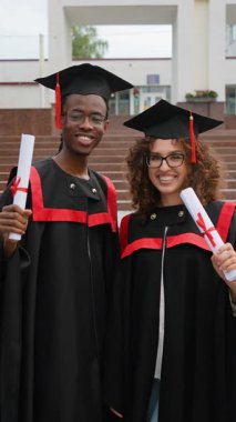 Mezuniyet elbisesi ve şapkalı iki farklı öğrenci akademik başarılarını kutluyor, mezuniyet töreninden sonra diplomalarını üniversite merdivenlerinin önünde tutarken mutlu bir şekilde gülümsüyorlar.