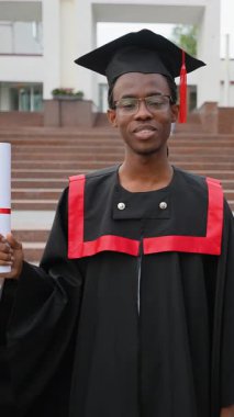 Genç Afro-Amerikan erkek, kep ve cübbeyle üniversite diplomasını elinde tutarak mezun oldu. Mezuniyet gününde akademik başarısını kutlayan gururlu ve gülümseyen bir öğrenci.