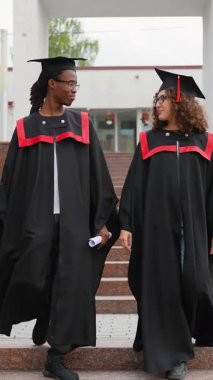 Geleneksel akademik önlüklü ve şapkalı mutlu çift üniversite merdivenlerinden aşağı iniyor, diplomalarını tutuyor, konuşuyor ve başarılı mezuniyet törenlerinden sonra gülümsüyor.