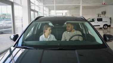 Muhtemel alıcılar yeni bir otomobilin içinde bir araba galerisinde otururken satıcı kadın da araçların özelliklerini sunarak adamın son satın alma kararını vermesine yardım ediyor.